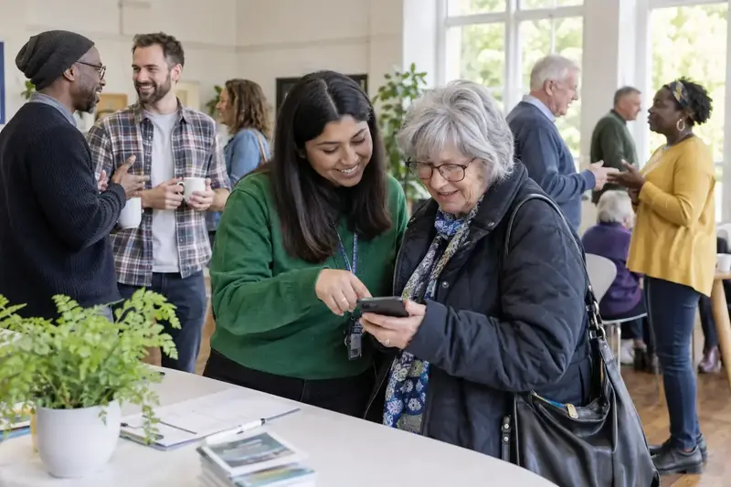 A community gathering in a bright hall, one member helping another sign in on a phone