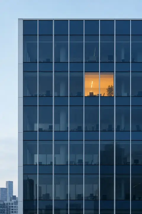 A navy glass office facade at night with a single window lit orange