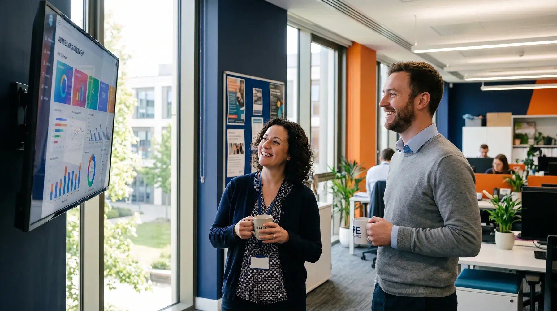 Two colleagues smiling at a dashboard on an office wall